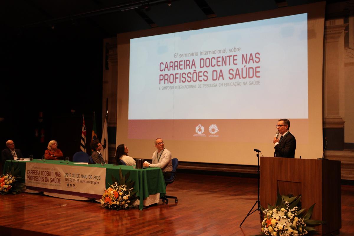 Mesa com toalha verde, ladeada por 5 adultos e, à direita, homem em pé com microfone na mão. Todos olham para telão com palavras sobre carreira docente nas profissões da saúde